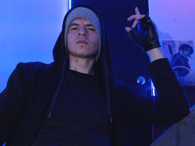 DreamHunterdz, met een grijze muts, een zwarte hoodie en handschoenen zonder vingers, maakt een "stil" gebaar met hun rechterhand, onder blauw licht..