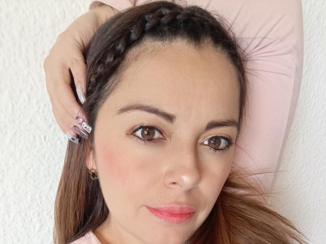 Seher's face, brown eyes, pink lips, light skin, braided brown hair, holding her head, pink shirt, white textured wall, pink nails with glitter, slight smile, relaxed expression.