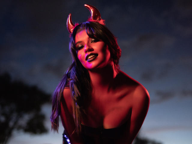 Katherine, com chifres vermelhos de diabo, maquiagem roxa e rosa no rosto, batom escuro e cabelos longos e ondulados, vestindo uma blusa sem alças, brilhando em luz roxa e vermelha..