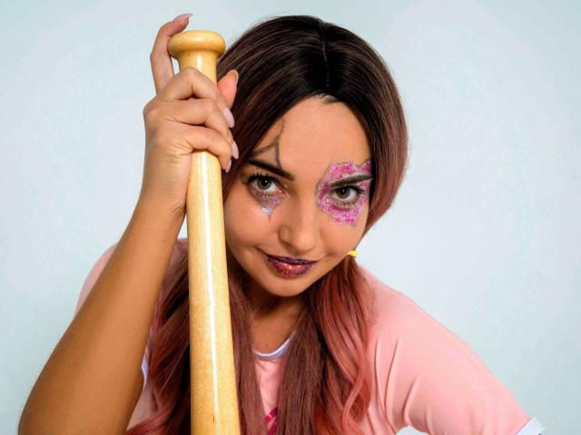 LettieSweet with pink eyeshadow, holding a wooden baseball bat, wearing a pink shirt, and a yellow earpiece, smiling at the camera. Her tits are partially visible.