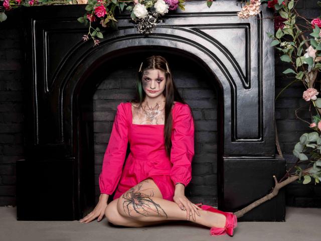 Nicole, mit langen braunen Haaren, einem pinken Kleid und Tattoos an ihrem Oberschenkel, sitzt vor einem schwarzen Kamin, trägt pinke Schuhe und ist von Blumen und Ranken umgeben..