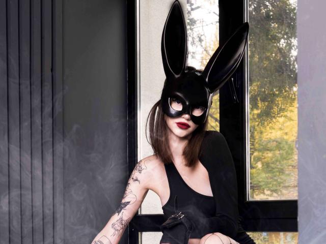SophieKitty with black rabbit mask, red lipstick, black tank top, tattooed arm, and smoky effect, sitting by window. Boobs are covered by tank top.