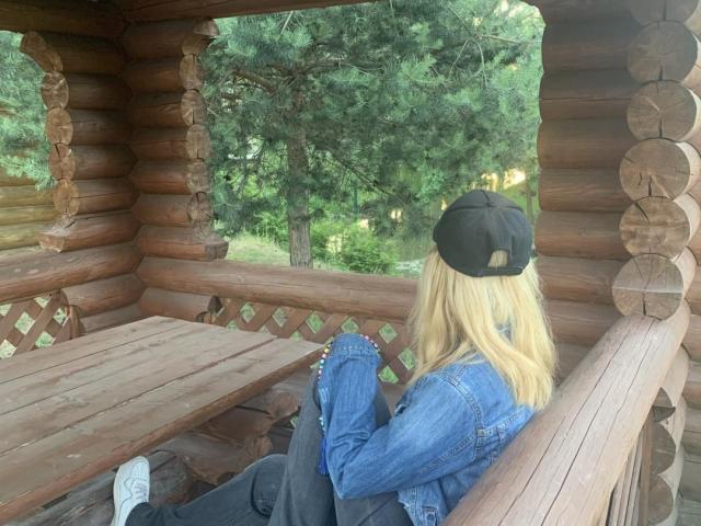 Tserera mit blonden Haaren und einer schwarzen Mütze sitzt auf einer Holzveranda, trägt eine Jeansjacke und eine schwarze Hose und hält ein Telefon. Ihr Hintern ist leicht sichtbar..