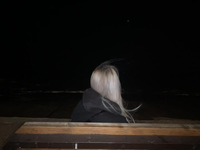 Blondine Tserera mit langen Haaren, in einer schwarzen Jacke, sitzt nachts auf einer hölzernen Bank und blickt auf einen winzigen, fernen, sichtbaren Stern im dunklen Himmel..