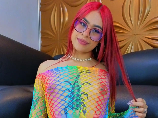 JazeVans, con i capelli rossi, gli occhiali e una collana di perle, indossa una maglia a rete arcobaleno che mette in mostra i suoi piccoli seni e i capezzoli piercing. Sorride, tenendosi i capelli..