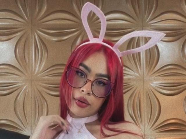 A JazeVans, com cabelo ruivo, orelhas de coelho cor-de-rosa e óculos redondos, tem um rosto fofo e está de língua para fora. Ela está usando um colar branco..