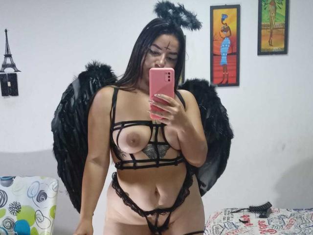 AbbyShiine with black wings and halo, wearing strappy black lingerie, showing her tits and pussy, holding a pink phone, standing in a bedroom, looking at the camera.