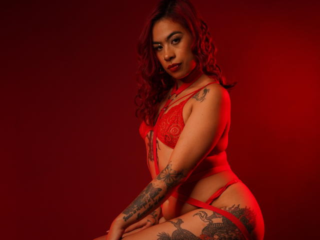 JazeVans con lencería roja, cabello rizado, tatuajes en los brazos y el muslo, sentada, luz roja, mirando a la cámara, mostrando los pechos, el trasero y la vulva, sexy, segura de sí misma, fondo rojo..