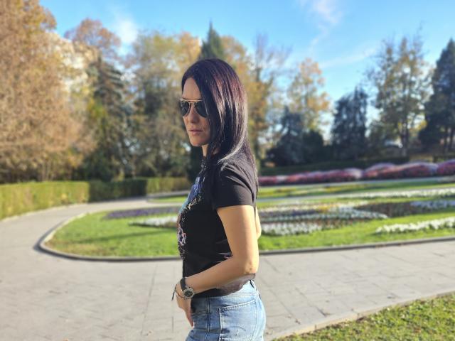 JoliDanielle with dark hair, wearing sunglasses, black t-shirt, and blue jeans, standing on a path, looking sideways, hands on waist, green garden in background, sunny day.