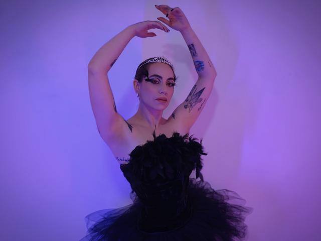 LuciaPerth, con los brazos levantados, vestida con un vestido negro con plumas, una tiara y tatuajes en los brazos. La iluminación morada y azul resalta su piel pálida y su maquillaje oscuro. Se ven tatuajes en el pecho..