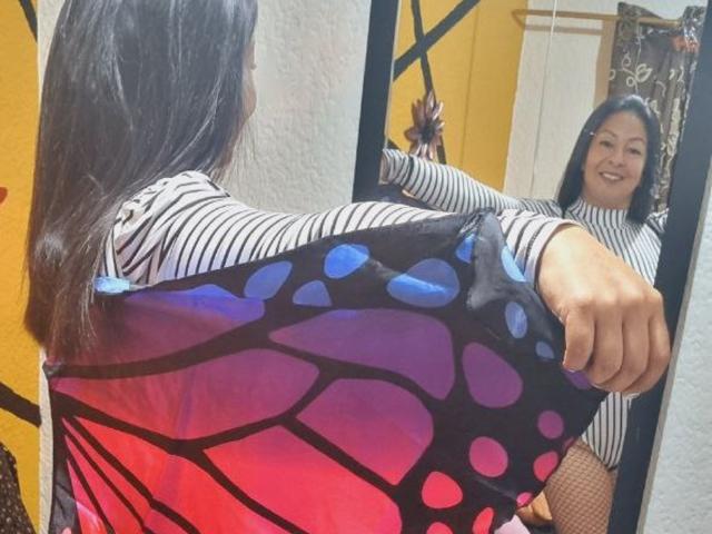 CatalinaJH con cabello negro largo, camisa a rayas, sosteniendo un tejido con estampado de mariposas, sonriendo a su reflejo en el espejo, diseño de mariposas en azul y morado, medias de red, sonriendo, con confianza, en un ambiente informal..