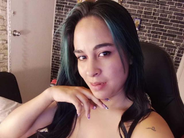 Krystal Brown, avec ses cheveux bleu turquoise, ses racines foncées, ses lèvres pulpeuses et un tatouage sur son épaule droite, pose sa main sur son menton, montrant ses doigts peints en violet..