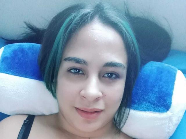 Krystal Brown, con cabello verde y negro, ojos azules y piel clara, acostada sobre un cojín azul y blanco, sonriendo, con una correa negra, mostrando su nariz y labios..