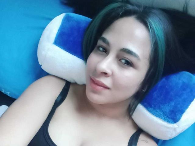 Krystal Brown, con cabello de color verde, camiseta negra, acostada sobre un cojín azul y blanco, sonriendo a la cámara, mostrando su linda cara y sus pequeños senos..