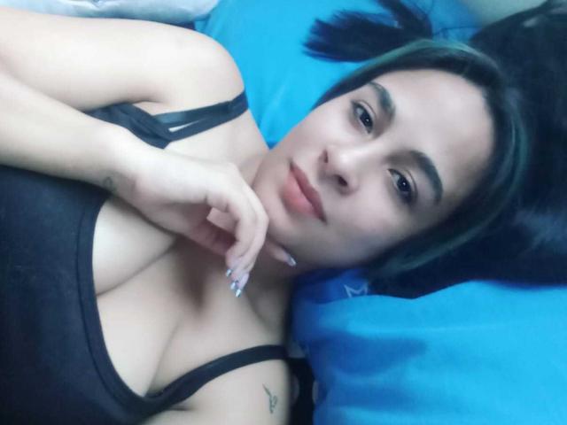 KrystalBrown with black hair, wearing a black tank top, lying on a blue pillow, showing her tits, hand near her chin, tattoo on her left arm, looking at camera.