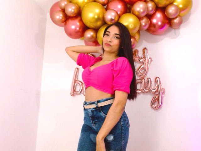 SarahyJhones mit langen, dunklen Haaren, einem pinken Crop-Top, das ihre Brüste zeigt, und blauen, hochgeschnittenen Jeans steht vor einer Wand aus pinken, goldenen und roten Luftballons. Sie schaut in die Kamera..
