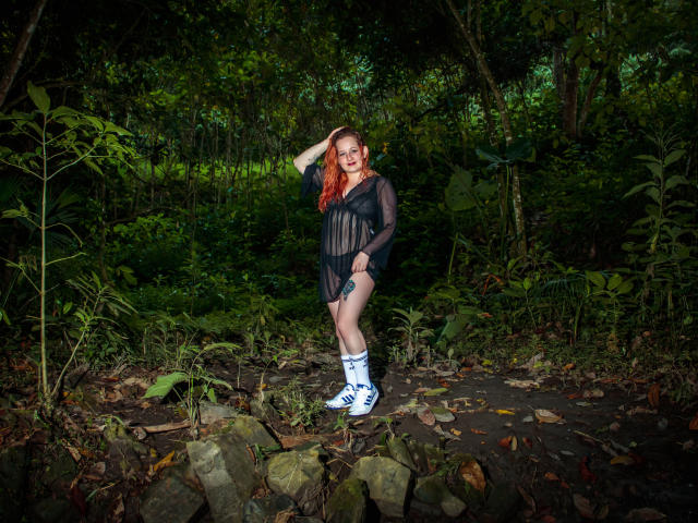 VioletaMora with red hair, black sheer dress, white sneakers, and knee socks, standing in a forest, hand on head, showing her tits and ass.