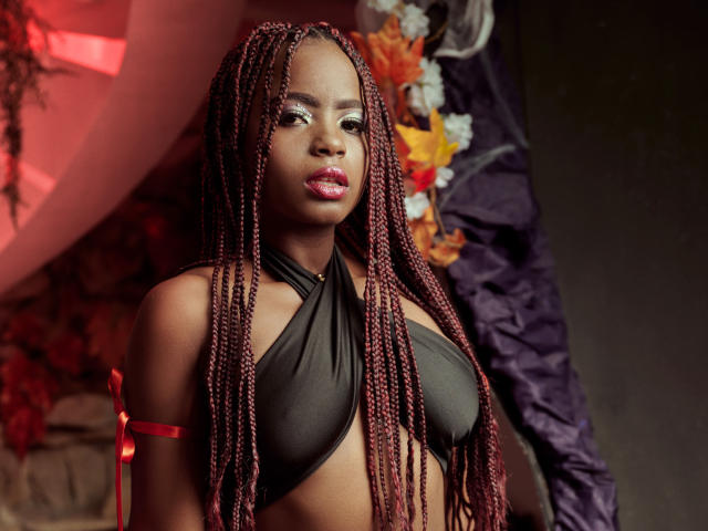 LindaBird with dark skin, red braided hair, wearing a black halter top, exposing her tits, red lipstick, and a red armband, looking sexy in a dimly lit room.