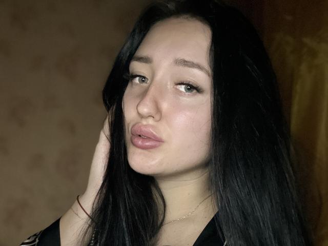 VishenKamur ha lunghi capelli neri, pelle chiara e labbra piene di colore rosa. Sta guardando la fotocamera con un'espressione leggermente corrucciata, e la sua mano destra tocca la testa..
