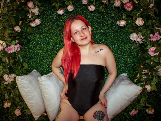 VioletaMora with red hair, black strapless leotard, tattoos, smiling, sitting on white pillows, surrounded by pink roses and green foliage, hands on thighs.