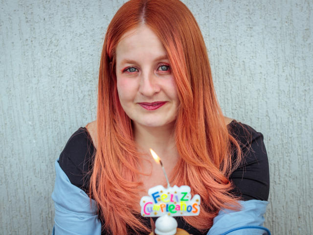 VioletaMora with red hair, black and blue shirt, holding a candle with a colorful text, smiling, blue eyes, slight pink lipstick, textured gray wall in background.