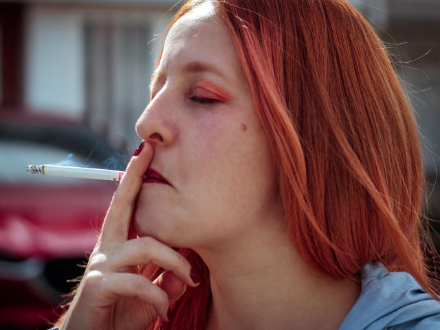 VioletaMora with red hair, holding a cigarette to her slightly parted lips, eyes half-closed, fingers resting on her chin, smoke curling up, wearing a blue shirt.