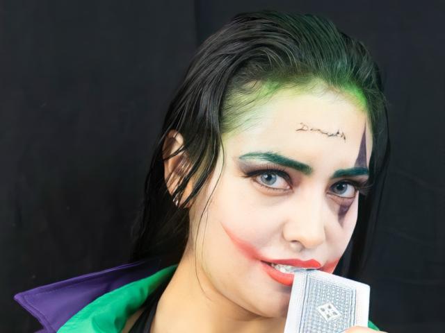 SilvannaReyes com maquiagem verde e preta, batom vermelho, cabelo preto, segurando um cartão, jaqueta verde e roxa, tatuagem verde na testa, sombra verde e um piercing no nariz..