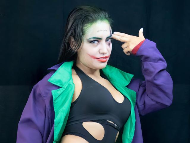 SilvannaReyes met een groene en paarse jas, een zwarte top met uitsparingen, make-up zoals de Joker, wijzend naar haar voorhoofd met een vinger, nat zwart haar, rode lippenstift en groene oogschaduw..