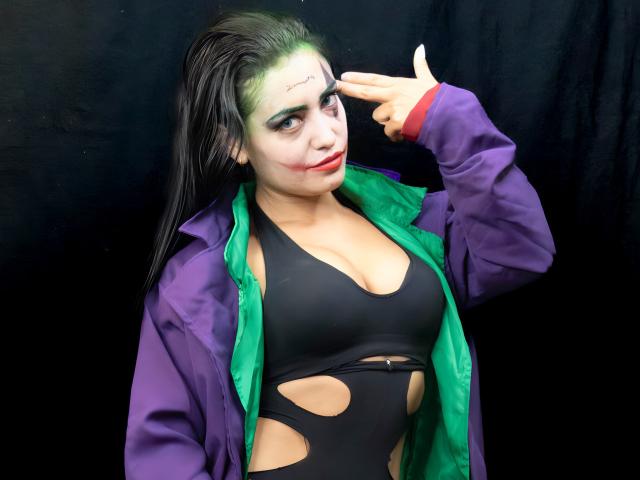 Silvanna Reyes met groene en zwarte make-up, een paars jasje met groene voering en een zwart topje met uitsparingen, wijst met een vinger naar haar voorhoofd en laat rode vlekken op haar wang zien..