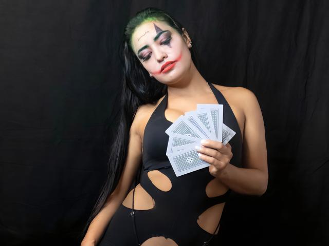 Silvanna Reyes met groen haar, witte gezichtsbeschildering, rode lipstick, een zwart topje met uitsparingen, die een stapel kaarten vasthoudt, en naar beneden kijkt. Op de achtergrond is zwart te zien. De borsten zijn zichtbaar door het topje..