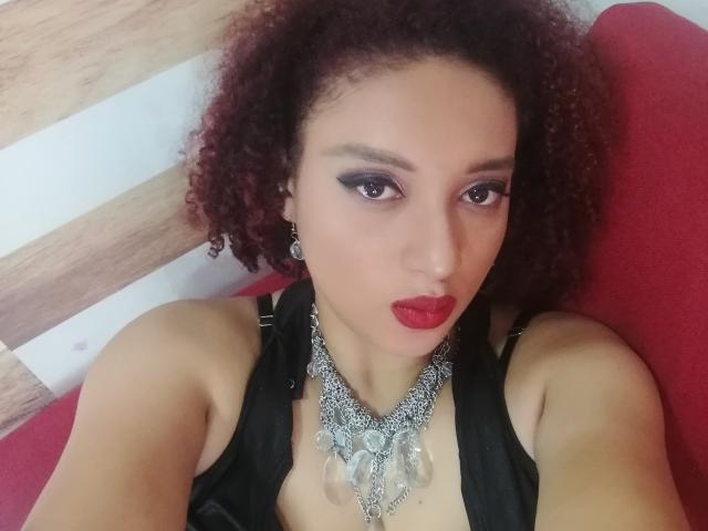 VioletaSwitch, con cabello rojo-marrón rizado, lápiz labial rojo, delineador de ojos negro, vestida con una blusa negra y un collar de plata grueso con un colgante grande, sentada en una silla roja..