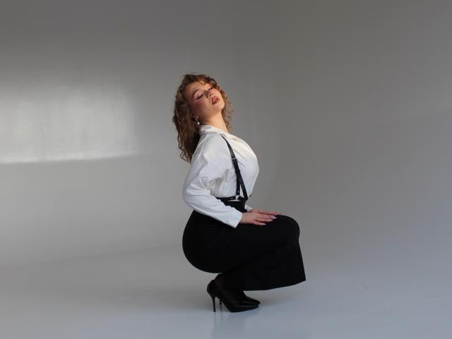 Stacy Yellig è in posizione di squat, indossa una camicia bianca, bretelle nere e pantaloni neri a vita alta, con tacchi neri, guarda in alto, con le mani sulle ginocchia, ha i capelli castani ricci e un leggero sorriso..