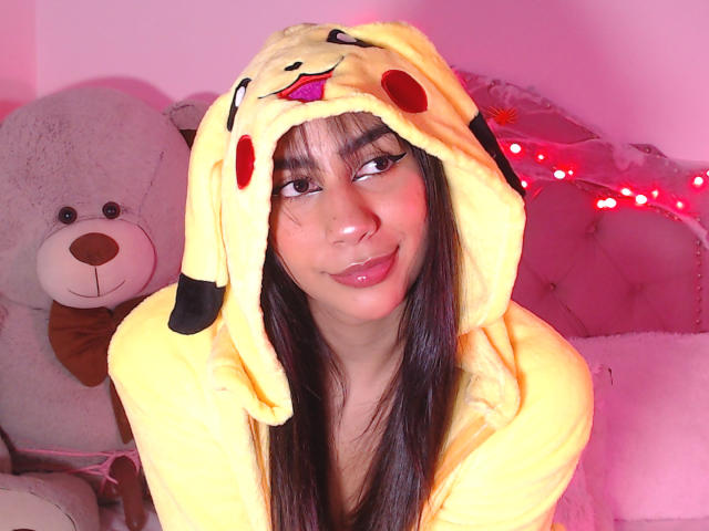 Jessica Soler, com longos cabelos pretos, vestindo um moletom amarelo do Pikachu, com lábios rosados delicados, sentada em frente a um grande ursinho de pelúcia branco, em um quarto rosa, com luzes de Natal vermelhas..