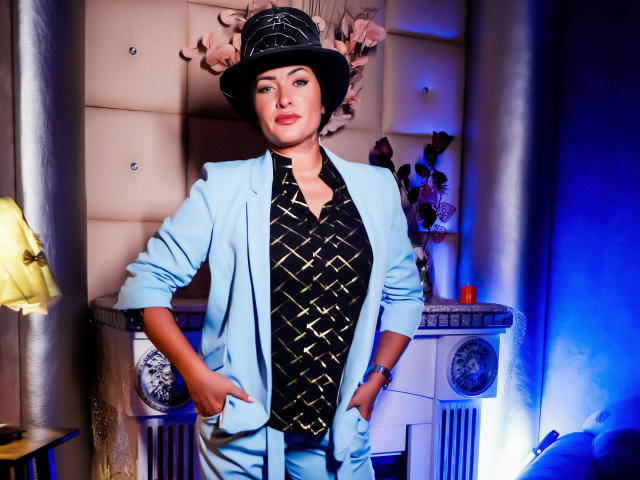 Anelisa Reys, con le mani in tasca, indossa un completo azzurro chiaro, una camicia nera a fantasia e un cappello nero con piume rosa, ed è in piedi con sicurezza in una stanza illuminata di blu..