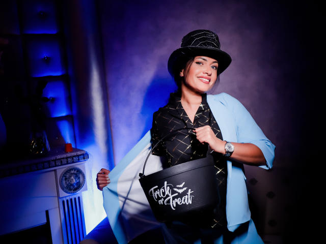 AnelisaReys, avec des seins, portant une veste bleu clair, un chapeau noir et une écharpe noire à motifs, tenant un sac noir pour les bonbons d'Halloween, souriant. Éclairage bleu et violet..