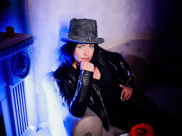 AnelisaReys mit blauen Haaren, einer schwarzen Lederjacke und einem Hut mit Schlangenmuster, die ihr Kinn in die Hand legt, verführerisch, blaues Licht, weißes Sofa, rote Blume unten rechts..