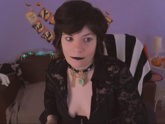 EverlyRays with black makeup, wearing a black lace top, necklace with green pendant, black choker, and exposed cleavage, sitting on a striped chair, in a purple room.