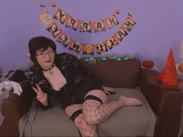 EverlyRays with pointy elf ears, black lace dress, fishnet stockings, and black lipstick, flashing a peace sign, sitting on a brown sofa, showing her ass. Halloween decorations behind.