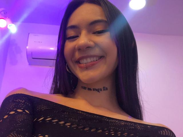 CieloSmith, avec de longs cheveux noirs, souriante, la langue tirée, portant un pull noir à épaules dénudées, un anneau au nez et un tatouage sous le menton, éclairage violet et blanc..