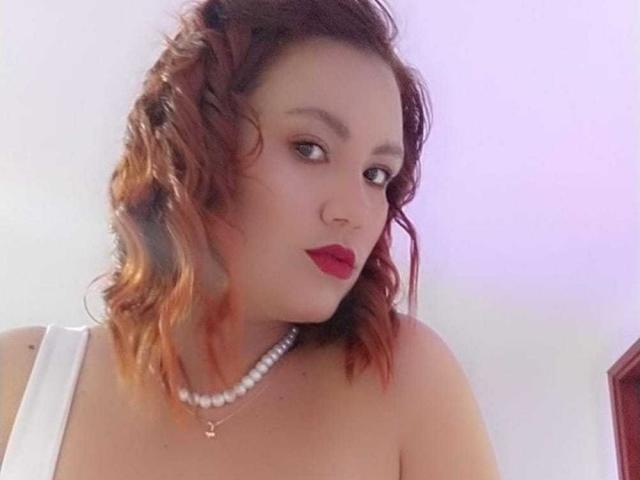 Sharon Grey mit lockigem, rotem Haar, die ein weißes Oberteil und eine Perlenkette trägt, hat roten Lippenstift und ein leichtes Lächeln und blickt zur Seite. Ihre Brüste sind teilweise sichtbar..