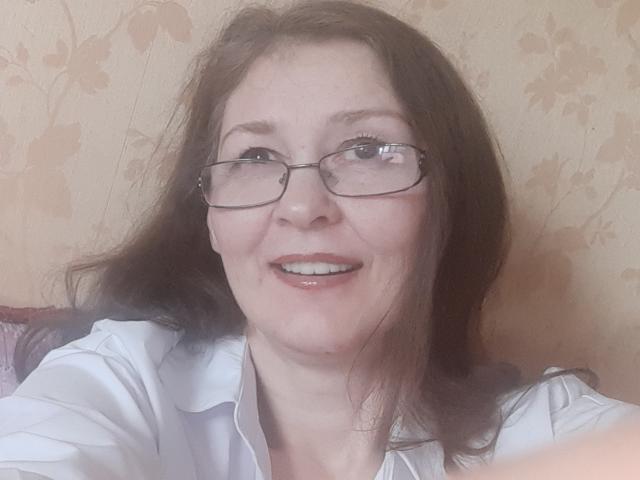 NellyMitchell with glasses, smiling, showing her teeth, wearing a white shirt, has brown hair, and fair skin. Her tits are partially visible under the shirt.