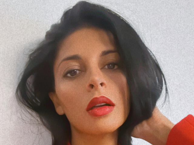 Rodalinda tem cabelos escuros e ondulados, olhos castanhos e lábios vermelhos. Ela está vestindo um suéter vermelho, com a mão no cabelo, olhando ligeiramente para o lado..