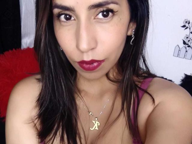 KatyKatt, con cabello castaño oscuro, lápiz labial rojo y un collar dorado con un dije en forma de la letra 'A', mirando a la cámara con ojos serios. Se ven los senos debajo de una blusa rosa..
