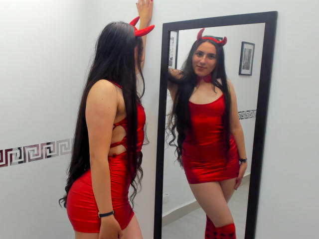 HelaViKy, con lunghi capelli neri, corna da diavolo rosse e un abito rosso lucido, mostra il suo sedere in uno specchio, in piedi con un braccio alzato, sorridendo al riflesso..