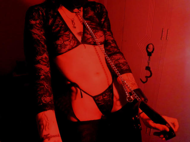 Envyv vestindo lingerie de renda preta, com uma corrente, algemas e uma tatuagem, segurando um brinquedo sexual preto, iluminada por luz vermelha, mostrando um abdômen plano e uma vulva pequena, coberta por renda..
