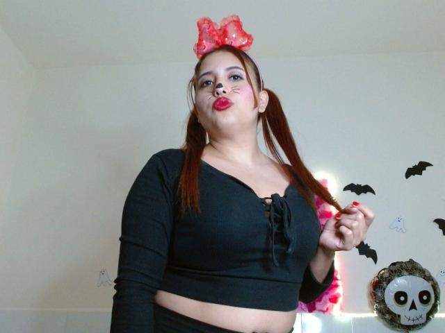 SamiLove com maria-chiquinhas, laço vermelho com bolinhas, cropped preto, mostrando os seios, lábios repuxados, segurando o cabelo, parede branca, morcegos de Halloween, adesivo de caveira..