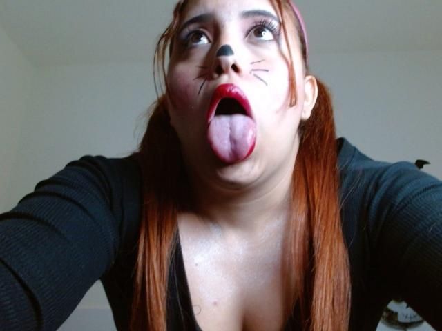 SamiLove, con lápiz labial rojo, pintura facial negra y la lengua afuera, tiene el cabello castaño en trenzas, una blusa negra y escote visible, en un selfie tomado desde un ángulo bajo..