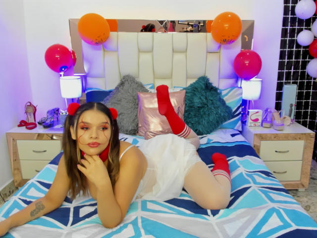 ValerieStone with red socks, white dress, and red hair bows, laying on a blue-patterned bed, showing her ass, surrounded by orange and red balloons, with a cute expression.