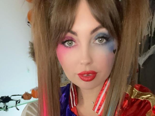 NinaTender with brown pigtails, pink and blue eye makeup, red lipstick, and a Harley Quinn costume, showing her face and upper chest. Boobs and pussy implied by costume.