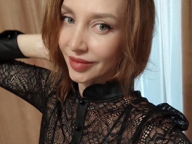 SimoniGrace ha i capelli castano chiaro, gli occhi verdi e il rossetto rosso. Indossa una maglia nera in pizzo con un colletto nero lucido, sorride leggermente con una mano dietro la testa..